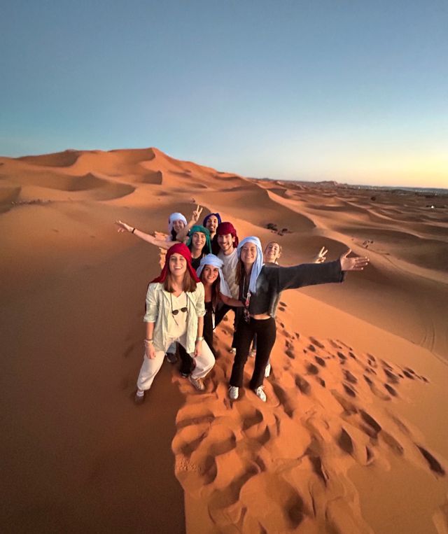 Eine WeRoad Gruppenreise posiert in der Abenddämmerung für ein Foto auf dem Kamm einer großen Sanddüne in der Wüste.