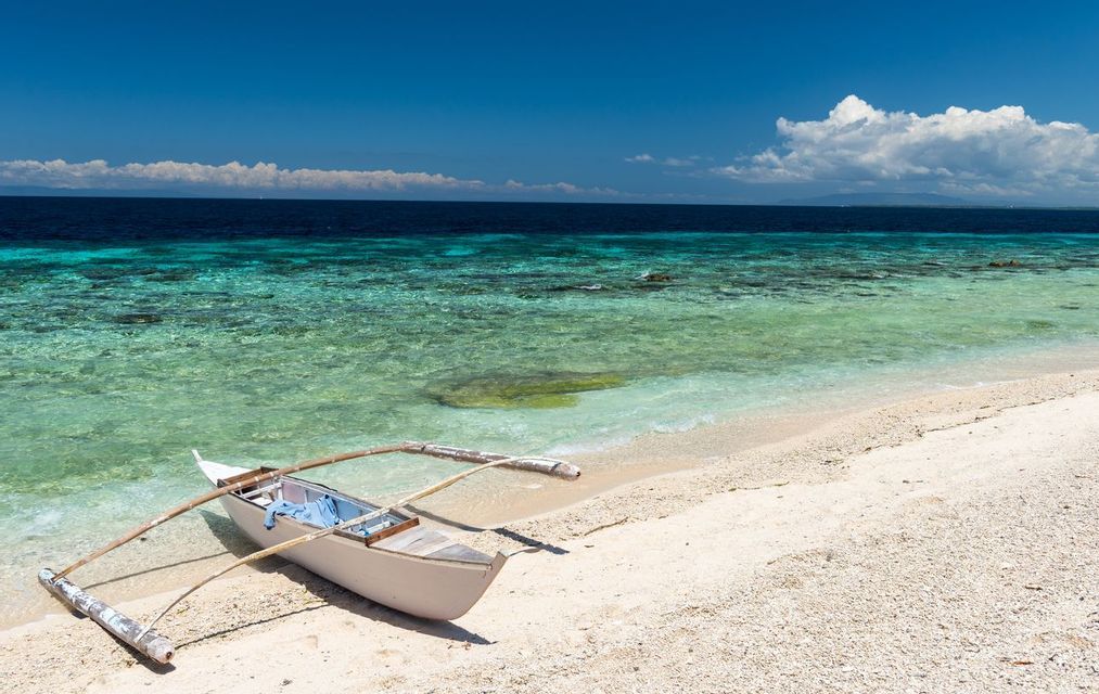 Una pequeña canoa de balancín descansa en una playa de arena blanca junto a agua clara y turquesa bajo un cielo azul brillante.