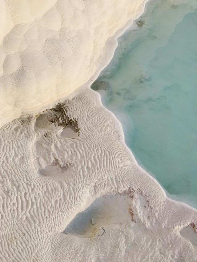 Una vista aerea di depositi minerali bianchi e testurizzati che formano terrazze accanto a uno specchio d'acqua calmo e turchese.