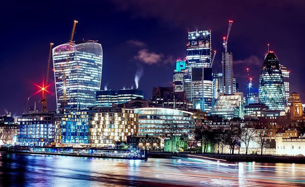 Uno skyline urbano di grattacieli ed edifici illuminati di notte, visto dall'altra sponda di un fiume con scie luminose colorate sull'acqua.