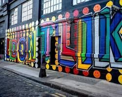 Graffiti colorati con lettere grandi e squadrate su un muro di un edificio, lungo una stretta via cittadina.