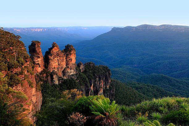 Tres grandes formaciones rocosas iluminadas por el sol se elevan desde un acantilado con vistas a un vasto valle montañoso cubierto de árboles bajo un cielo despejado.