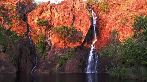 Dos cascadas caen por una pared de roca roja iluminada por el sol hacia un lago oscuro y tranquilo rodeado de árboles verdes.