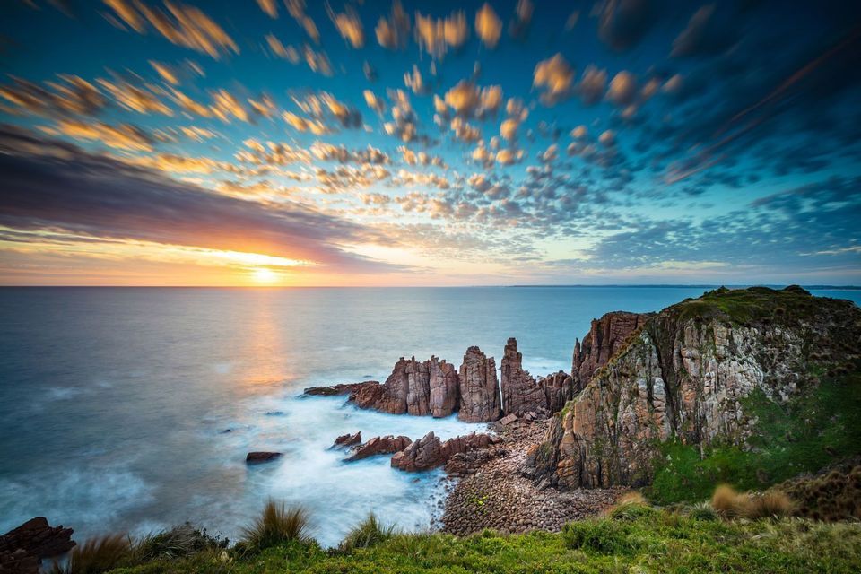 Jagged rock formations meet the ocean on a coastline under a colorful sunset sky with long-exposure clouds.