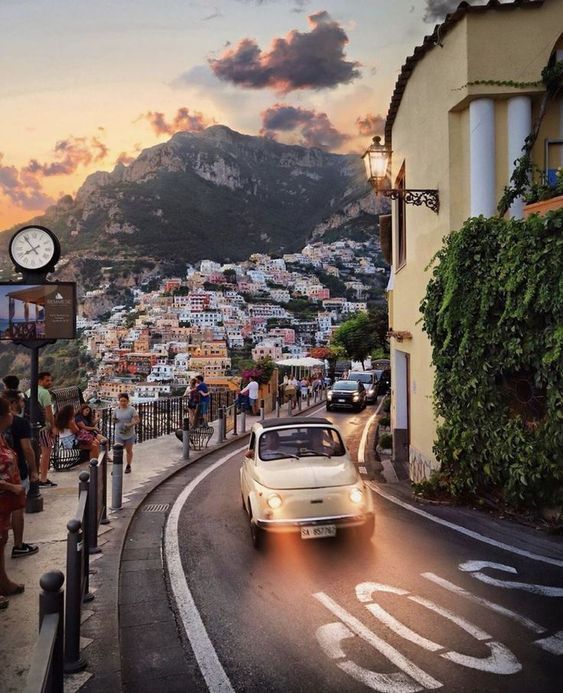 Un'auto d'epoca color crema con i fari accesi percorre una strada panoramica che domina una cittadina collinare al tramonto.