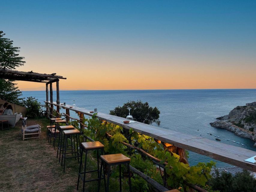 Un bar all'aperto con un lungo bancone in legno e sgabelli si affaccia sul mare e su una costa rocciosa al tramonto.