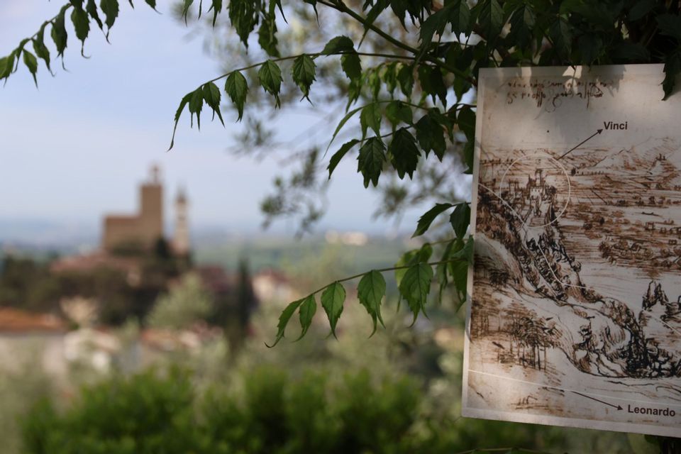 Una mappa di Vinci, opera di Leonardo, pende da un ramo d'albero, con il paese visibile sullo sfondo sfocato.
