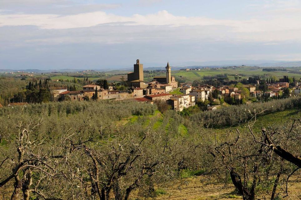 Un borgo storico con un'imponente torre in pietra su una collina, circondato da uliveti e dolci colline verdi sotto un cielo nuvoloso.