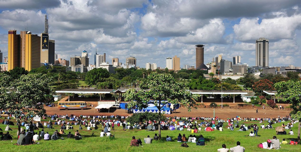 Una gran multitud de personas se sienta en el césped de un parque con un horizonte moderno de la ciudad visible al fondo bajo un cielo nublado.