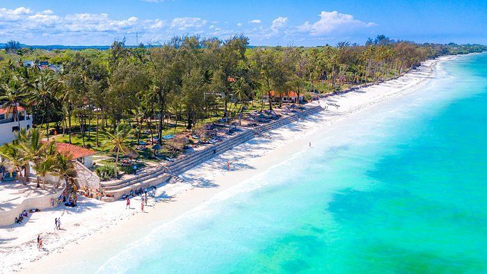 Vista aérea de una playa de arena blanca con agua turquesa, bordeada de palmeras y complejos turísticos bajo un cielo azul.