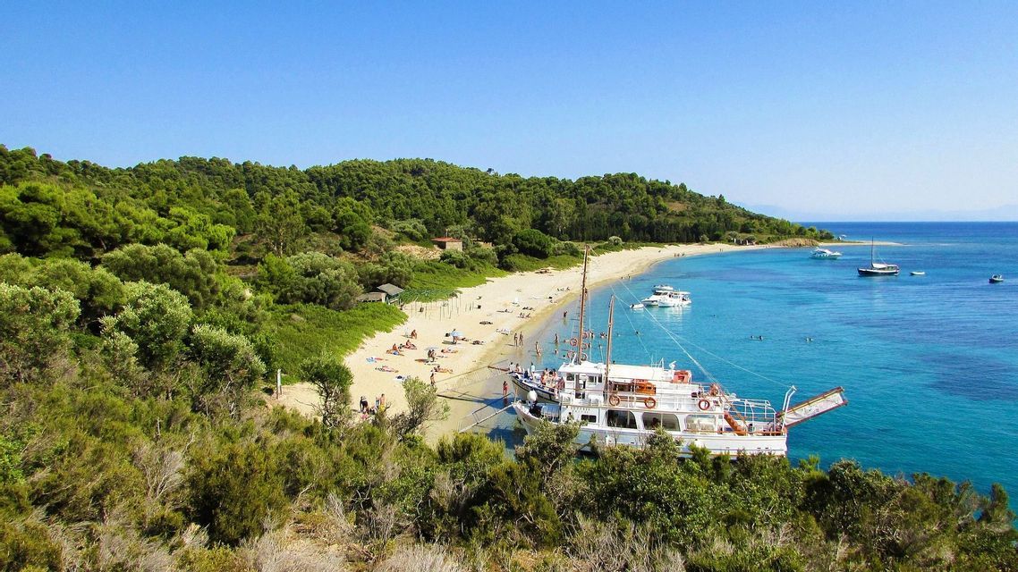 Una grande barca turistica bianca è ormeggiata in una baia turchese, accanto a una spiaggia sabbiosa con persone, con rigogliose colline verdi come sfondo sotto un cielo sereno.