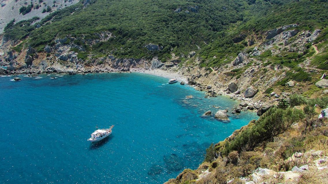 Una vista aerea di una caletta appartata con due barche che galleggiano in acqua turchese cristallina, accanto a una collina rocciosa e verde.