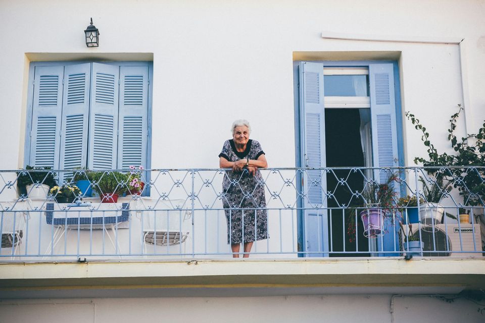 Una donna anziana con i capelli bianchi sorride appoggiata a una ringhiera del balcone davanti a un edificio bianco con persiane blu.