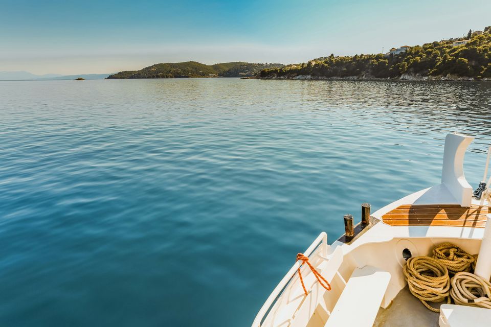 Prua di barca su acqua calma e azzurra, verso una costa verde e collinare sotto cielo limpido.