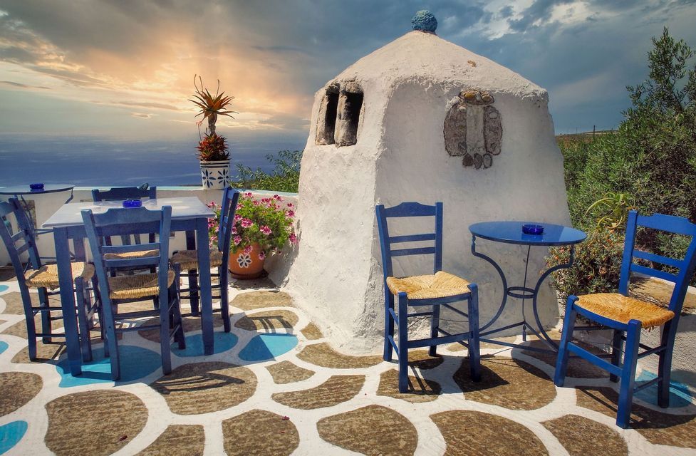 Tavoli e sedie blu su un patio in pietra affacciato sul mare, accanto a un edificio imbiancato a calce al tramonto.