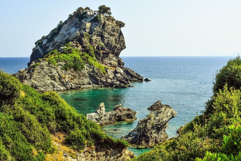 Una piccola cappella arroccata su un isolotto roccioso circondata da un mare turchese, con rigogliose scogliere verdi in primo piano.