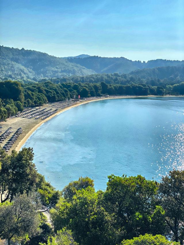 Vista dall'alto di una spiaggia sabbiosa che si estende lungo una tranquilla baia blu, con file di ombrelloni e colline boscose.