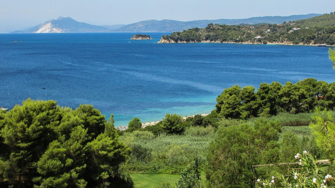 Una vista dall'alto di una baia costiera con acqua blu profonda, circondata da alberi verdi e montagne distanti sotto un cielo sereno.