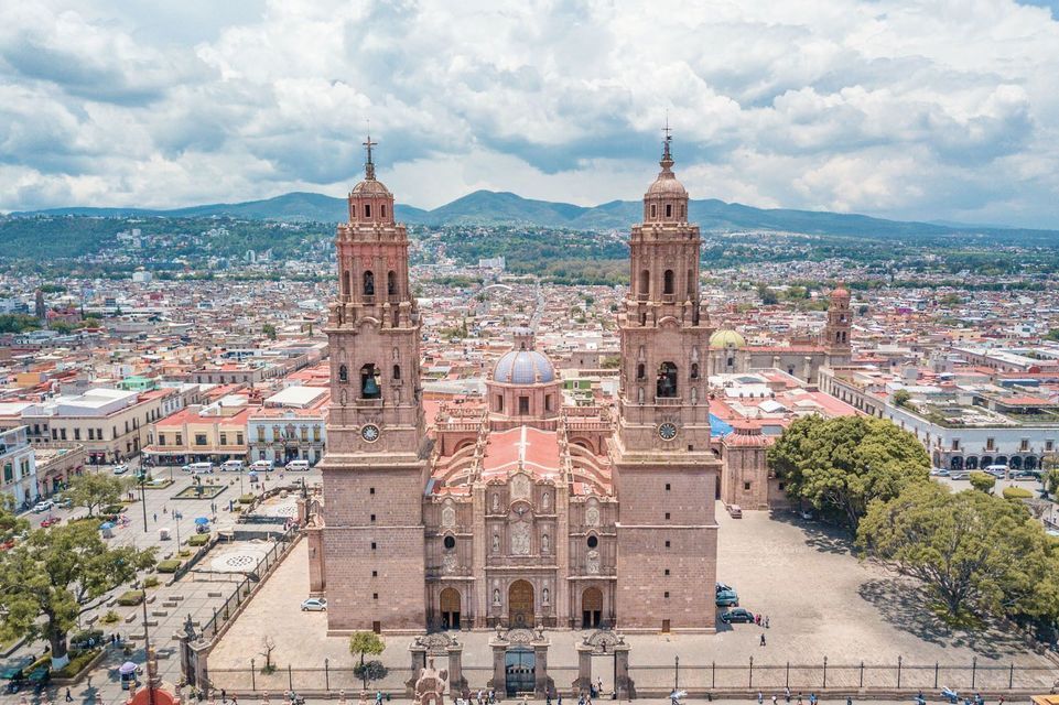 Vista aerea di un'ornata cattedrale in pietra con due grandi campanili, situata in una piazza di città e con le montagne sullo sfondo.