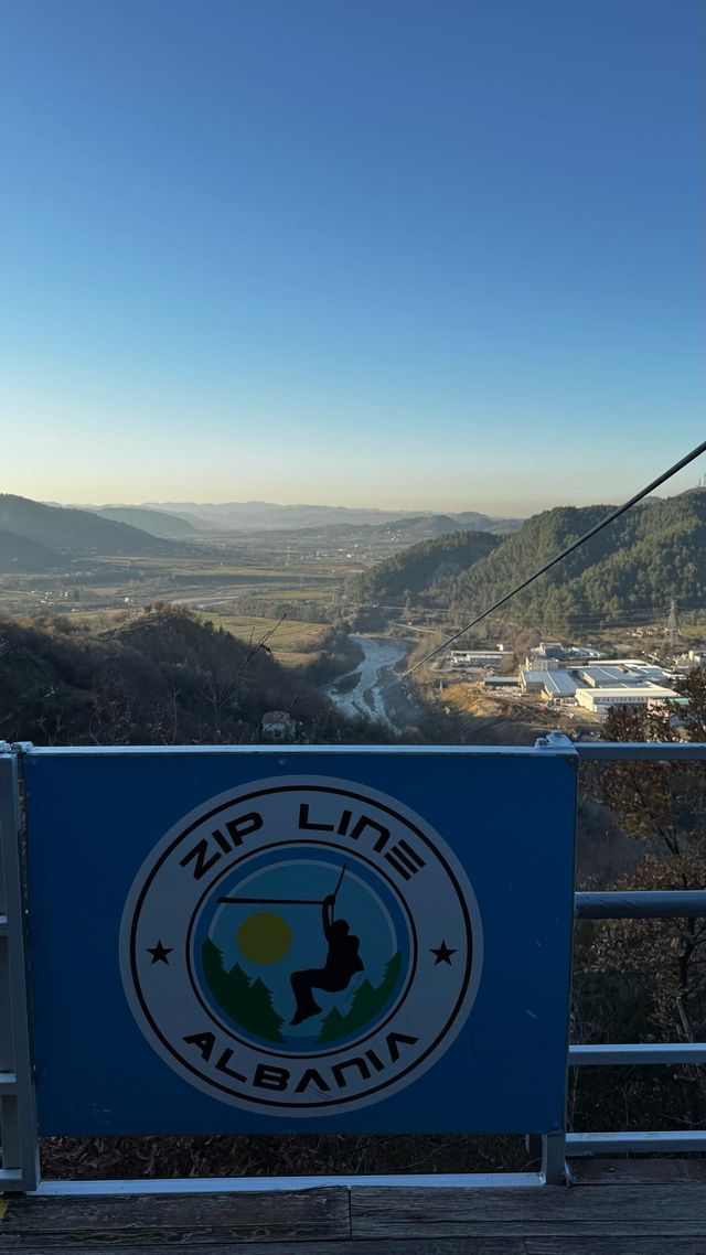 Un cartello blu 'Zip Line Albania' si affaccia su una vasta valle con un fiume e colline boscose sotto un cielo sereno.