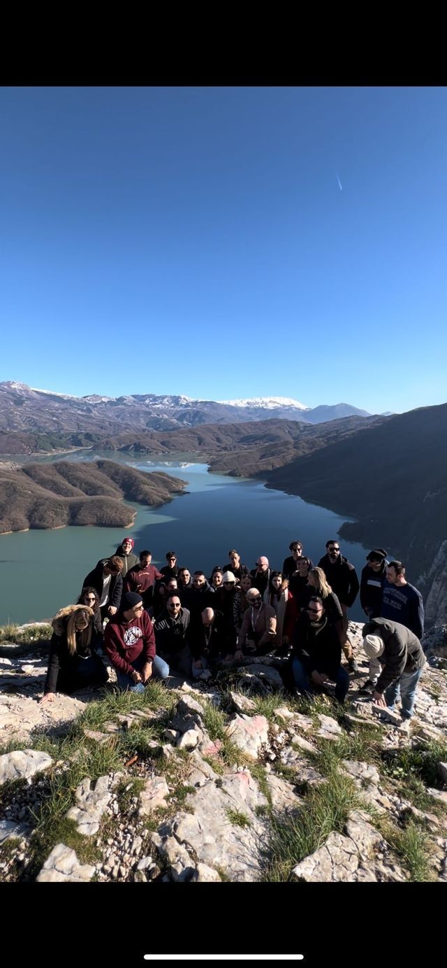 Un gruppo WeRoad in posa per una foto su una cima rocciosa che domina un lago panoramico e montagne innevate in lontananza.