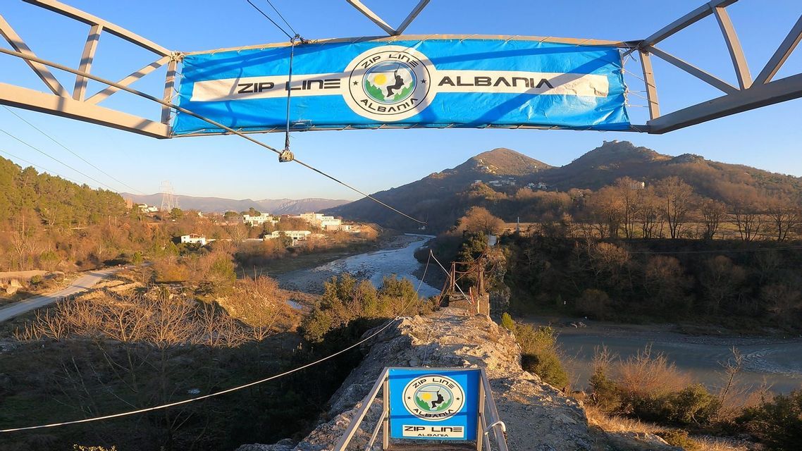 La piattaforma di partenza per 'Zip Line Albania' domina un'ampia valle fluviale con montagne sotto un cielo limpido e azzurro.