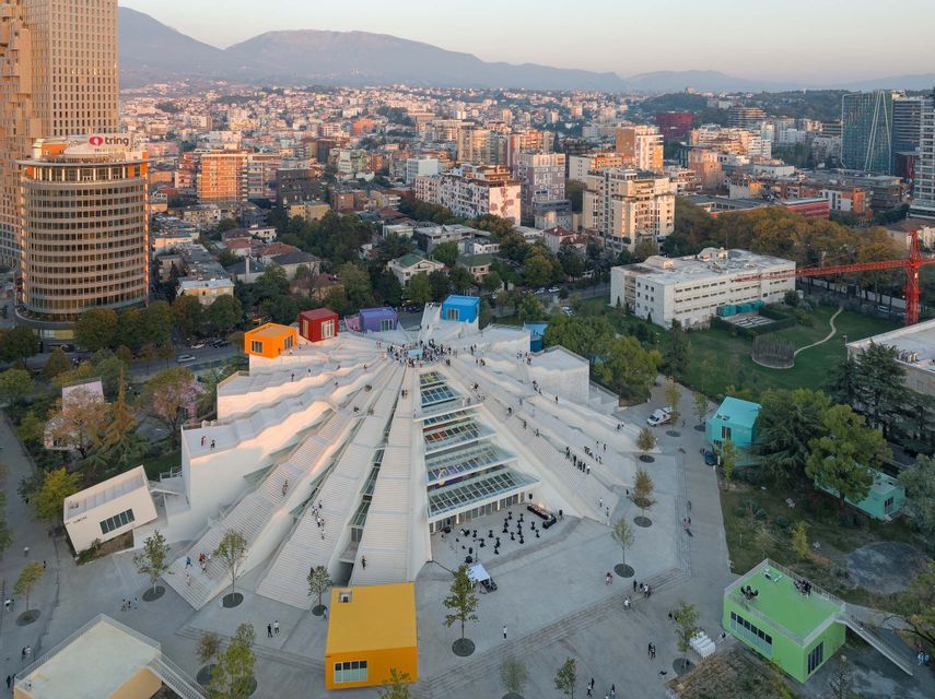 Una vista aerea di un moderno edificio bianco con terrazze calpestabili, immerso in un paesaggio urbano con montagne al tramonto.