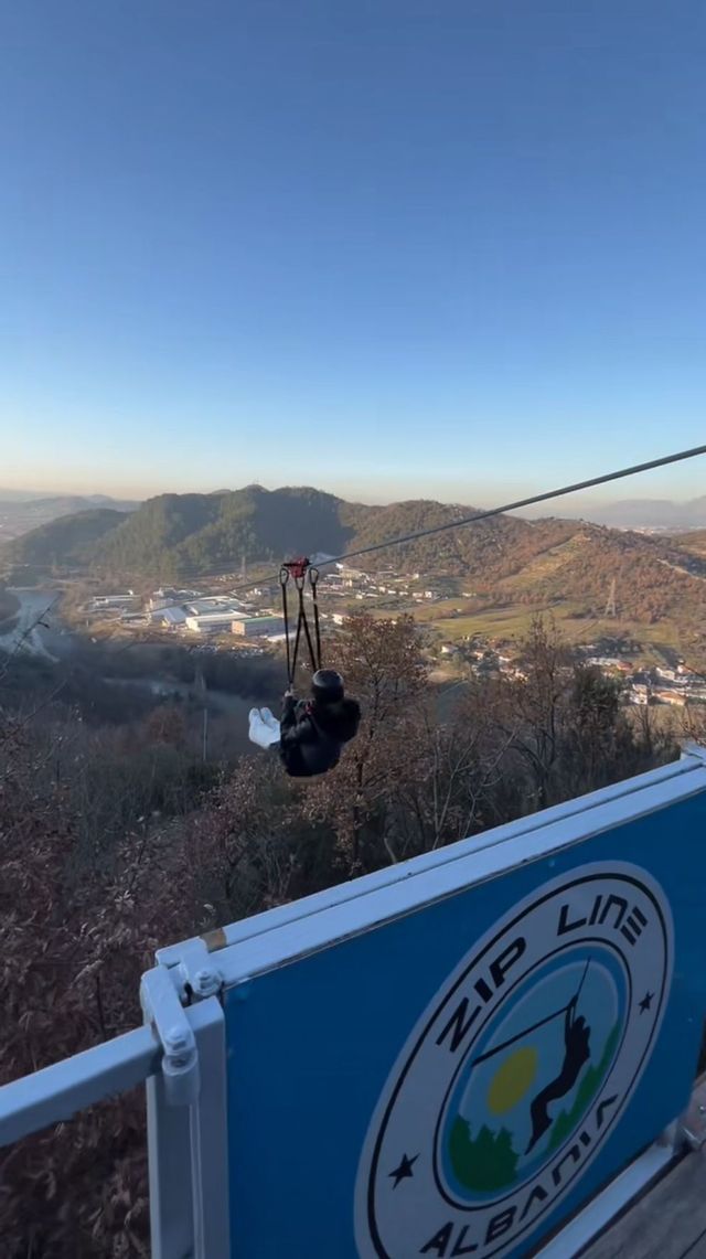 Una persona con un casco scivola su una zipline in alto sopra una valle con colline boscose e una città in lontananza.
