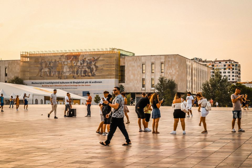 Folla di persone che camminano e sostano in un'ampia piazza urbana pavimentata, con edifici moderni sullo sfondo al tramonto.