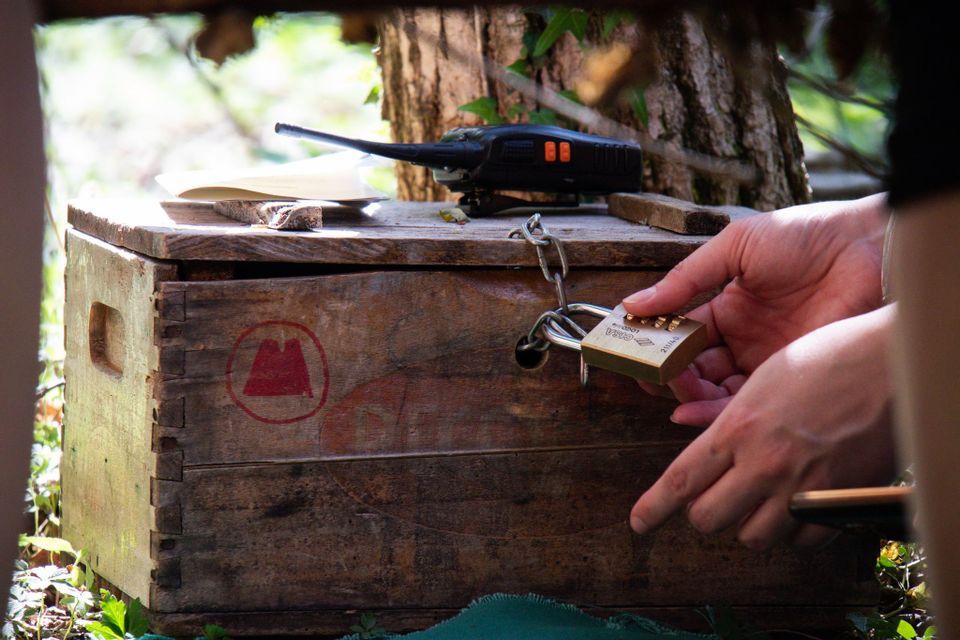 Le mani di una persona tentano di aprire un lucchetto a combinazione su una cassa di legno rustica, con un walkie-talkie appoggiato sopra.