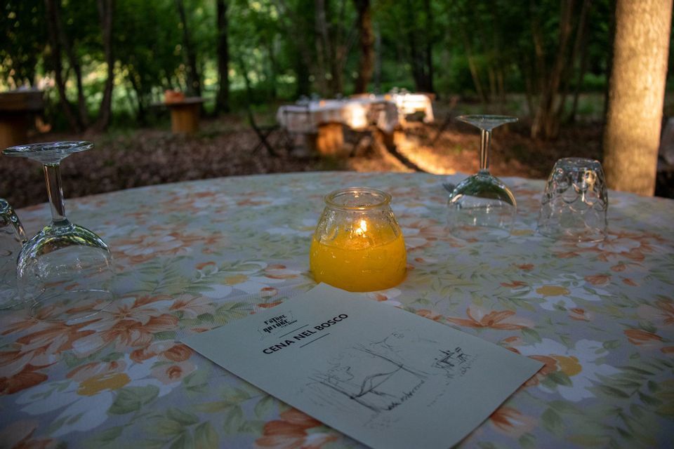 Una tavola apparecchiata per cena nel bosco, con bicchieri capovolti, una tovaglia floreale e una candela accesa in un barattolo.
