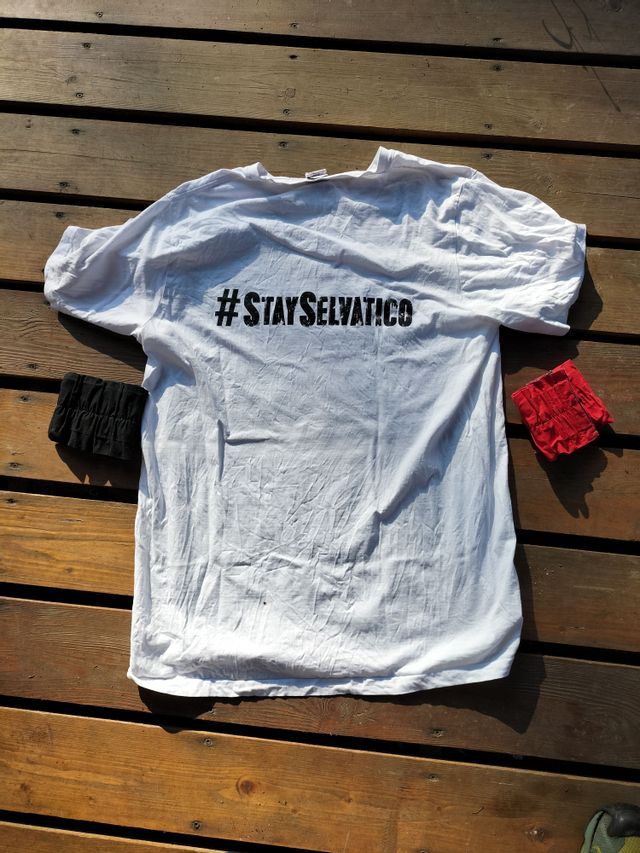 Una maglietta bianca con l'hashtag #STAYSELVATICO stampato in nero giace su un pontile di legno accanto a uno scrunchie rosso e uno nero.