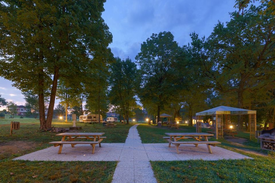 Tavoli da picnic e un sentiero pavimentato in un campeggio con alberi e roulotte, illuminati dalle luci al crepuscolo.