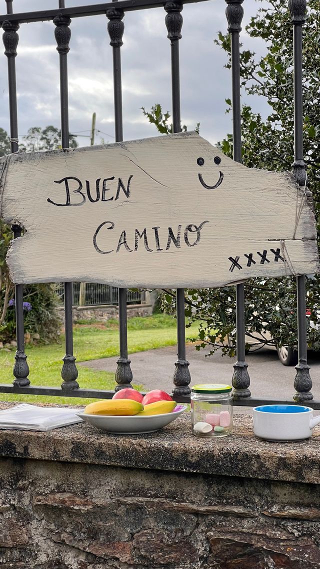 Un cartello di legno con la scritta 'Buen Camino' pende da una recinzione metallica sopra un muro di pietra con frutta e dolci.