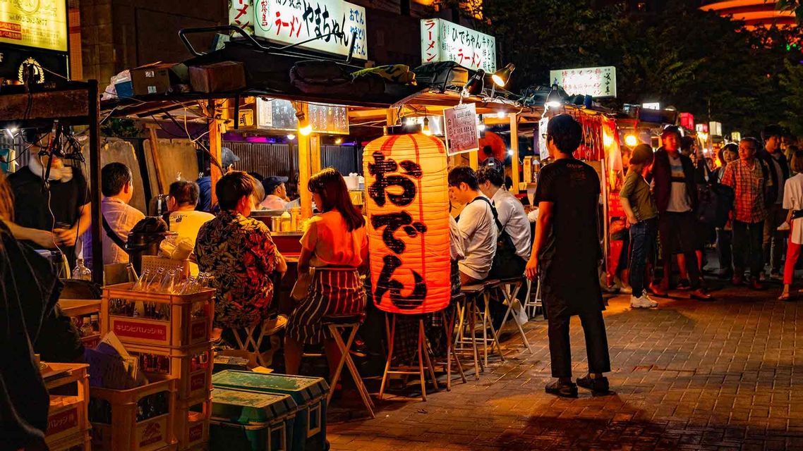 Una vivace bancarella di street food di notte, con avventori che mangiano al bancone accanto a una grande lanterna giapponese illuminata.
