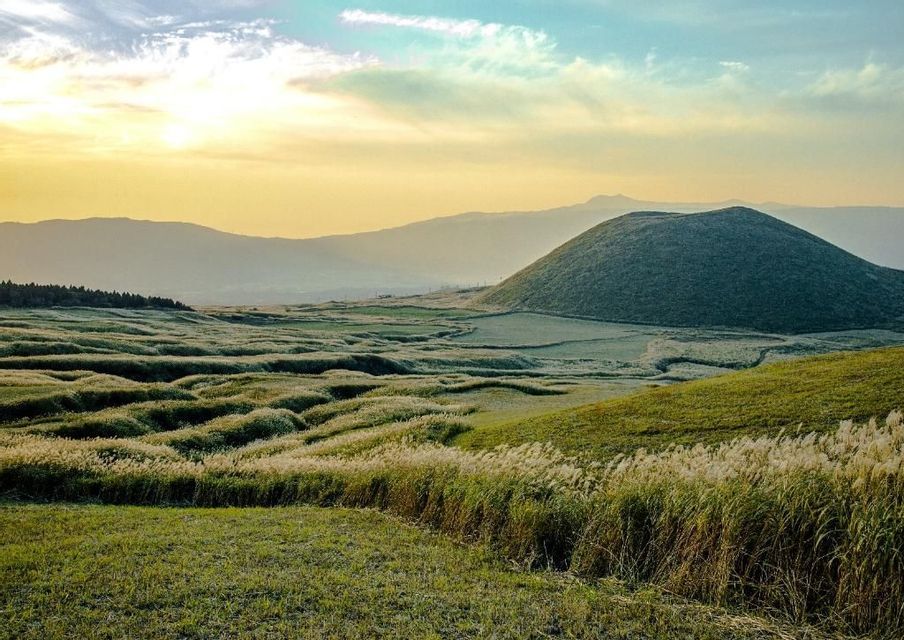 Un paesaggio di dolci colline verdi e un grande monte conico coperto di erba piumosa sotto un cielo velato e colorato.