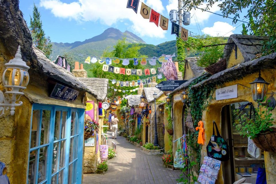 Una stretta strada acciottolata in un villaggio fantasy con negozi decorati per Halloween e una montagna visibile sullo sfondo.