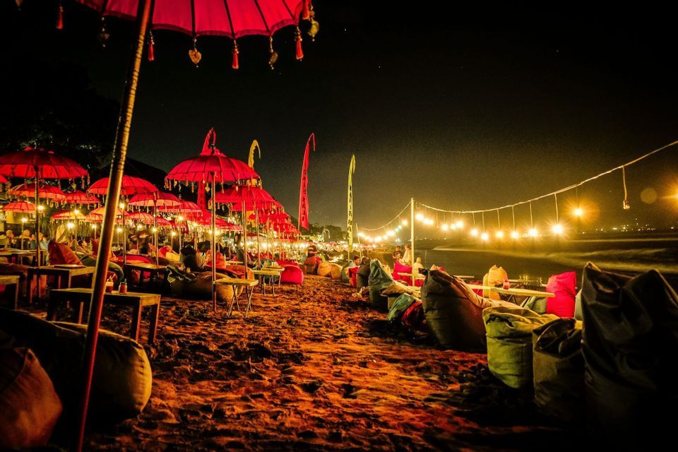 Fila di ombrelloni rossi e pouf su una spiaggia sabbiosa di notte, illuminati da calde lucine.