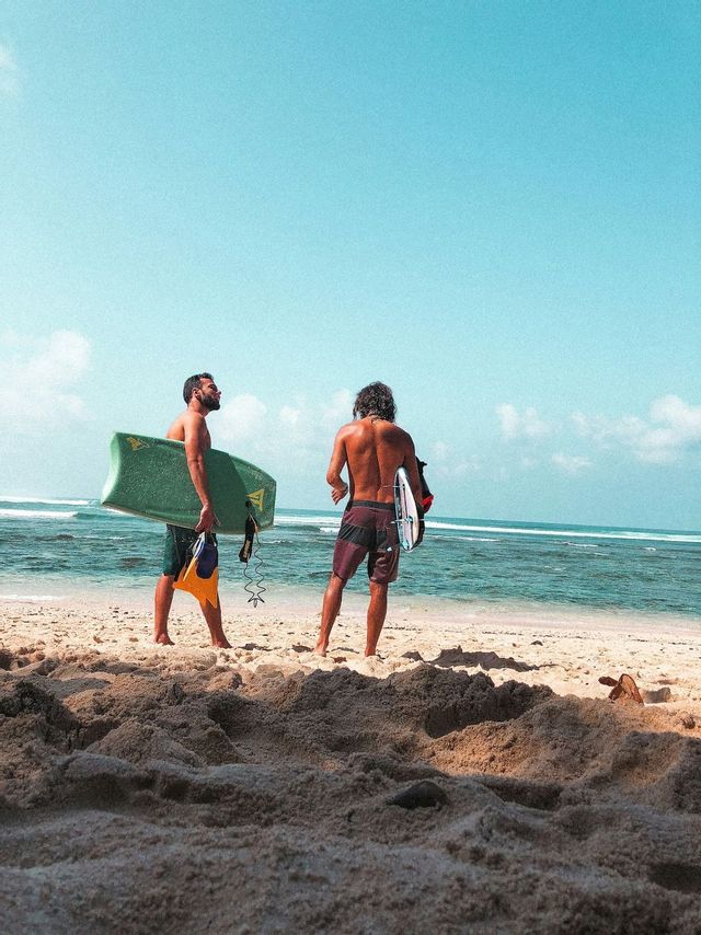 Due uomini di un viaggio di gruppo WeRoad si trovano su una spiaggia sabbiosa con bodyboard, guardando l'oceano sotto un cielo azzurro.