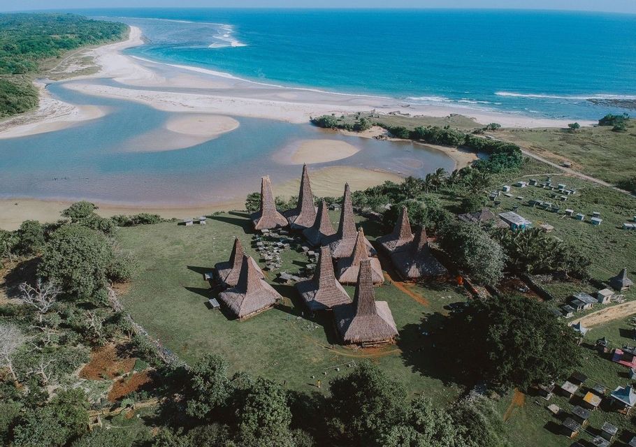 Una vista aerea di un villaggio tradizionale con alti tetti di paglia su una collina erbosa che si affaccia su una spiaggia, una laguna e l'oceano.