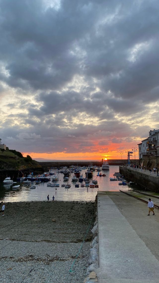 Un porto pieno di piccole barche al tramonto, con un sole arancione basso all'orizzonte sotto un cielo molto nuvoloso.