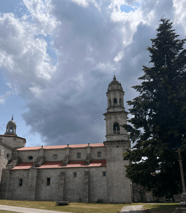 Una chiesa in pietra con campanile e tetto in tegole rosse si erge accanto a un alto albero sempreverde sotto un cielo drammatico e nuvoloso.