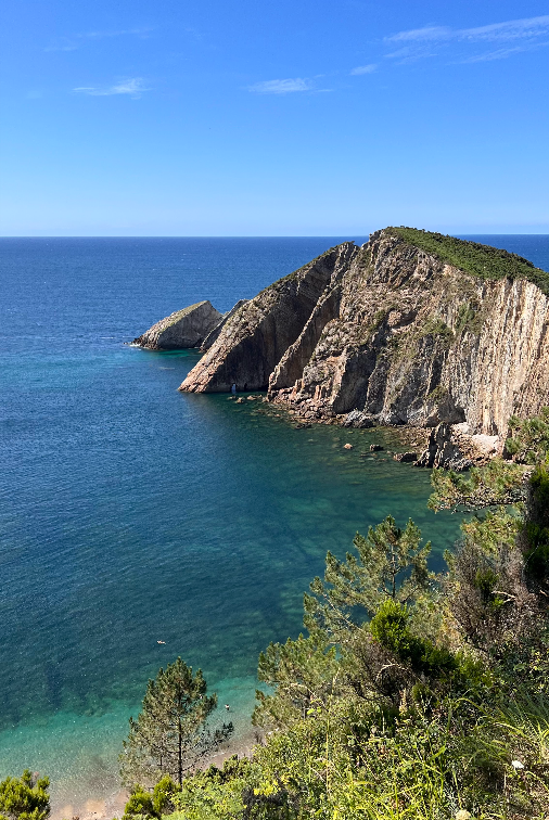 Una vista dall'alto di un promontorio roccioso che si estende in un mare calmo e turchese con una piccola caletta sottostante.