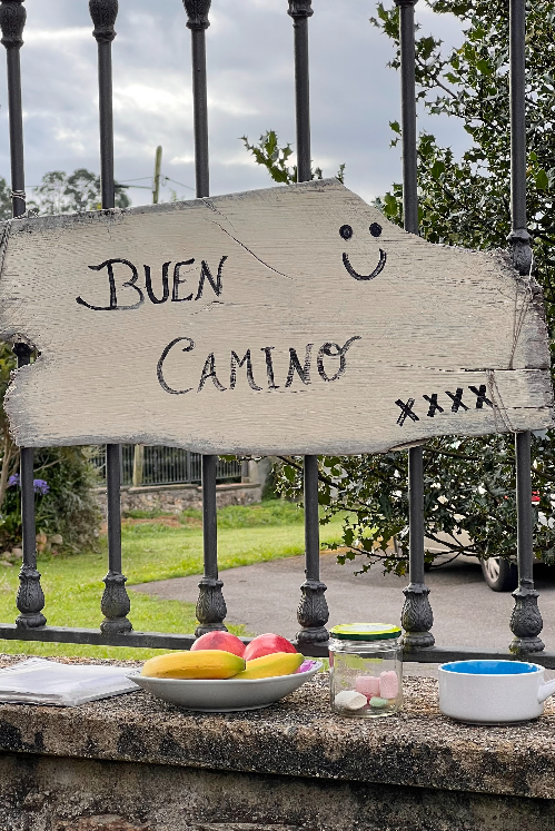 Un cartello di legno con la scritta 'Buen Camino' è appeso a una recinzione, sopra un muro di pietra che contiene una ciotola di frutta e un barattolo di dolci.