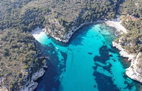 An aerial view of a secluded cove with turquoise water, a white sand beach, and surrounded by green, tree-covered cliffs.