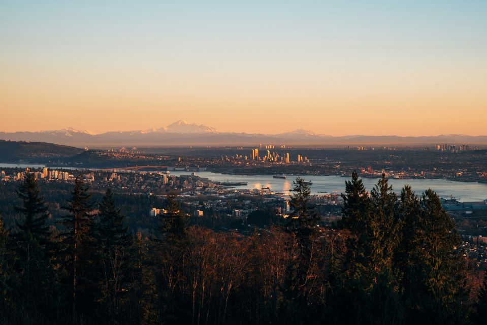 Vue plongeante sur une silhouette urbaine au bord de l'eau au lever du soleil, avec des montagnes enneigées au loin et la cime des arbres en contrebas.