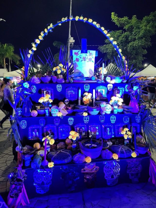 Un tradizionale altare ofrenda del Giorno dei Morti illuminato da luci blu di notte, decorato con teschi, fiori e foto incorniciate.