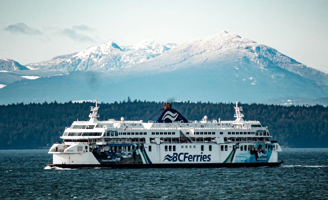 Un grand navire blanc de BC Ferries navigue sur l'eau, avec un rivage boisé et des montagnes enneigées en arrière-plan.