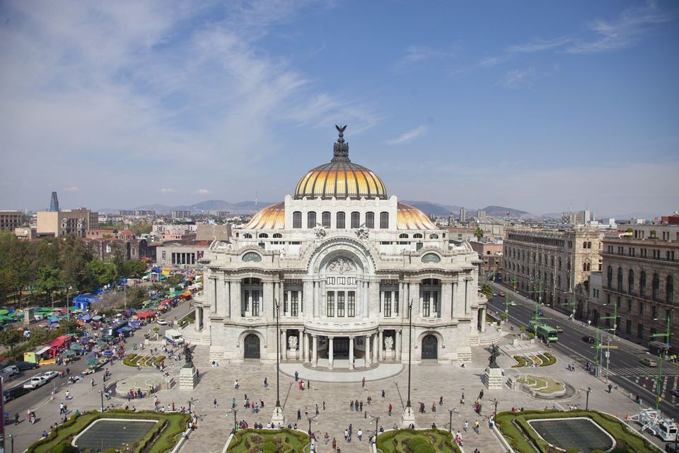 Un edificio blanco ornamentado con una cúpula dorada, centrado en una bulliciosa plaza de la ciudad bajo un cielo azul brillante, con un paisaje urbano de fondo.