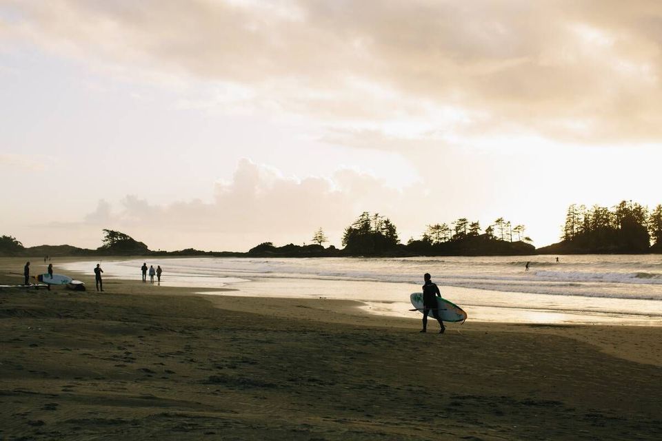 Une personne en combinaison de plongée porte une planche de surf le long d'une plage de sable au coucher du soleil, avec d'autres surfeurs en arrière-plan.